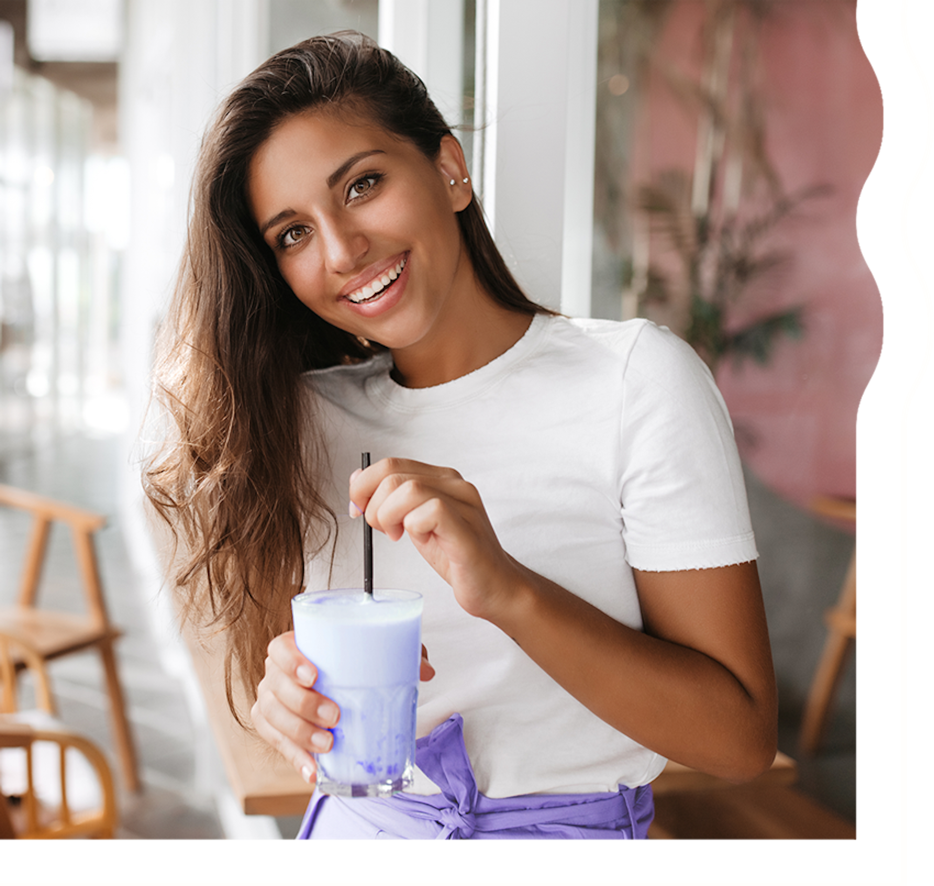 A woman with tan skin and long, dark brown hair is smiling and looking directly at the camera. She is holding a creamy, light blueish purple shake.