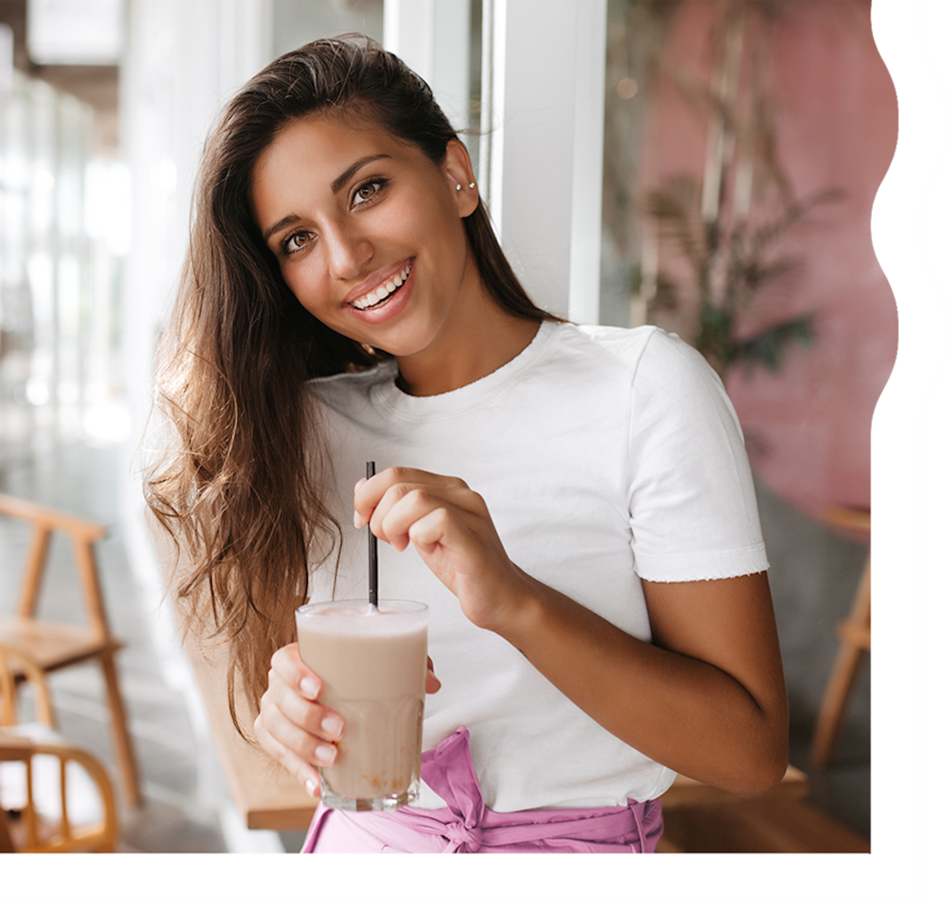 A woman with tan skin and long, dark brown hair is smiling and looking directly at the camera. She is holding a rich, chocolatey colored shake.