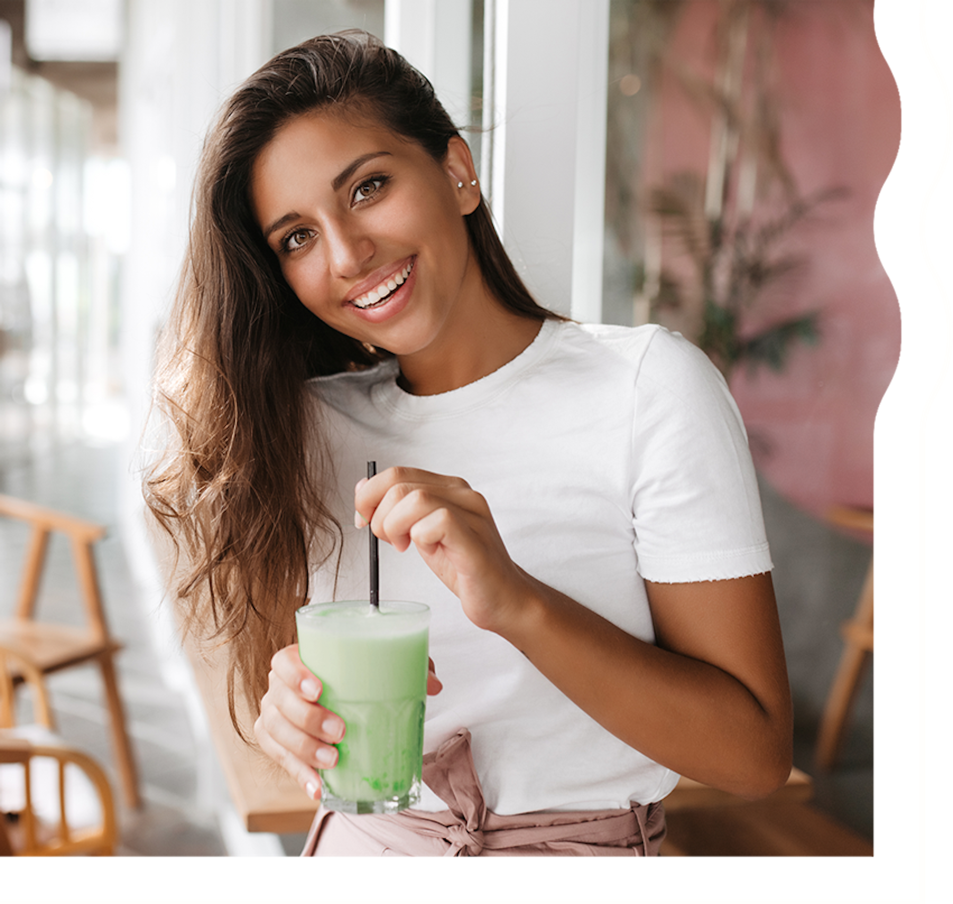 A woman with tan skin and long, dark brown hair is smiling and looking directly at the camera. She is holding a creamy, pistachio green shake.