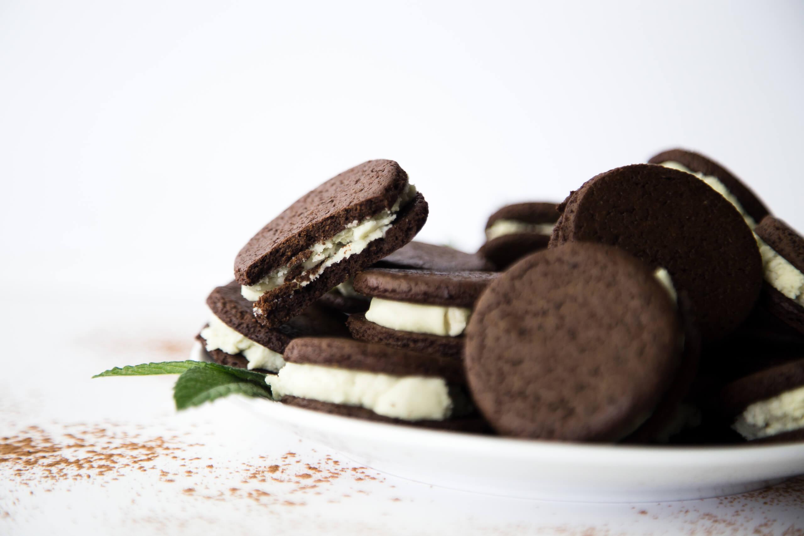 Pile of dark chocolate sandwich cookies with white cream filling on a white plate, accompanied by green mint leaves and scattered brown powder on a white surface.