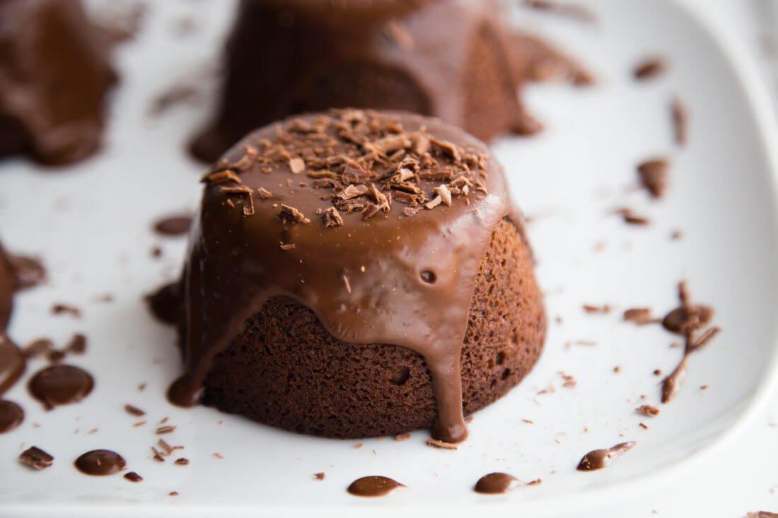 Chocolate cake with chocolate ganache and shavings on a white plate, with another cake blurred in the background.